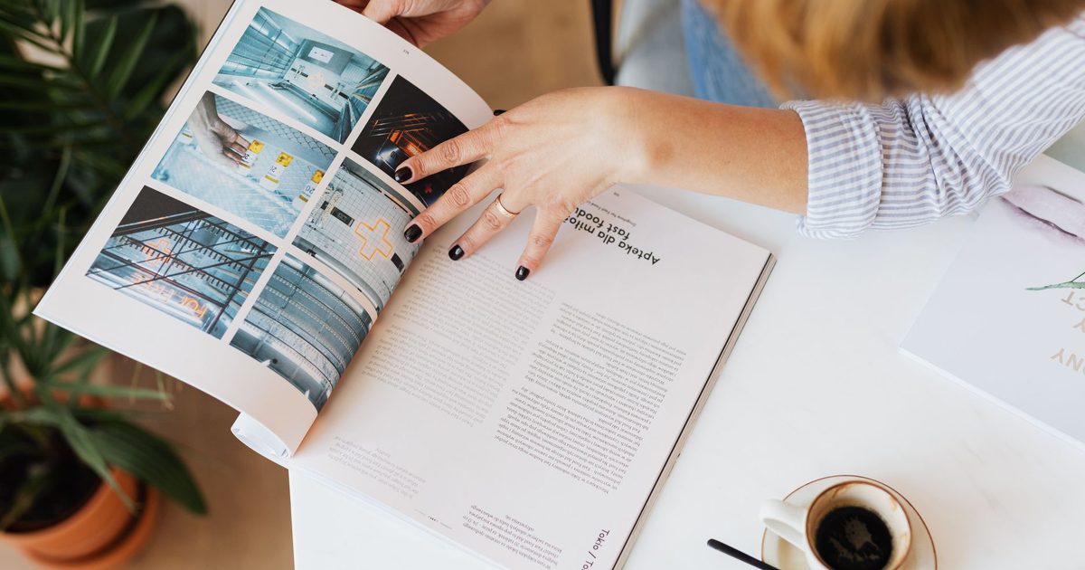 Mains feuilletant un magazine sur un bureau, à côté d’une tasse de café