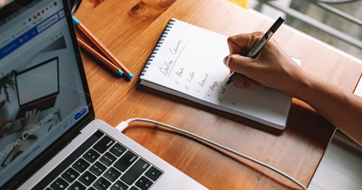 Main écrivant des notes sur un carnet à côté d’un ordinateur portable ouvert
